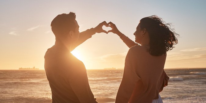Couple, sunset and heart sign at beach with hands for date, relationship and romantic moment together. Man, woman and love gesture by ocean with partner and back view for travel, holiday and loyalty.